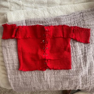 Röd offshoulder topp med snörning - Säljer en superfin röd offshoulder topp med snörning framtill. Den har en smickrande passform och volangdetaljer vid ärmslut och överkanten. Perfekt för festliga tillfällen eller en utekväll. Toppen är i bra skick och redo för nya äventyr! ❤️