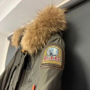 Grön jacka från Parajumpers - Säljer en grön parkajacka från Parajumpers Masterpiece Series. Jackan har en pälsfodrad huva och flera praktiska fickor. Perfekt för kalla vinterdagar med sin robusta design och varma foder. Den har en snygg patch på ärmen och dragkedja framtill. Passar perfekt för både vardag och utomhusaktiviteter. Bra skick.