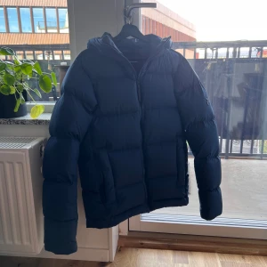 Mörkblå dunjacka från Peak Performance - En snygg och varm jacka med huva från Peak Performance i nyskick. Jackan har en dragkedja framtill och två fickor på sidorna.  Storlek S, passar även mindre M. Köptes för 4099kr, använd en gång sedan dess. Motsvarar dagens model ”Rivel Jacket”.