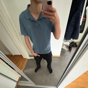 Blå pikétröja - Säljer en stilren blå pikétröja i normal passform. Perfekt för både vardag och lite finare tillfällen. Tröjan har korta ärmar och en klassisk krage med knappar. Skön och enkel att matcha med olika outfits.