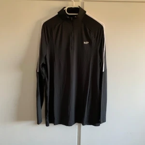 Svart långärmad träningströja från MP - Säljer en svart långärmad träningströja från MP i storlek S. Tröjan har en halv dragkedja och vita detaljer längs ärmarna. Perfekt för träning eller löpning, med en bekväm passform och stilren design. Passa på att köpa denna snygga och funktionella tröja! Helt ny originalförpackning följer med
