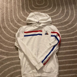 Vit hoodie från Le Coq Sportif - Säljer en snygg vit hoodie från Le Coq Sportif i storlek M. Den har coola röda och blå ränder över bröstet och längs ena ärmen. Perfekt för en sportig look eller bara för att chilla. Den är i bra skick och superbekväm!