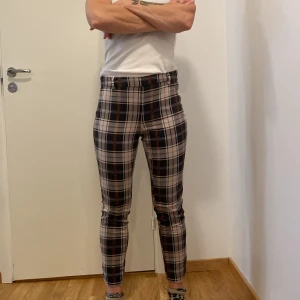 Rutig byxa från H&M - Säljer ett par snygga rutig byxor från H&M i storlek 38. Byxorna har en tight passform och är perfekt för höst och vår. De har ett klassiskt rutmönster i svart, beige och orange. Superbekväma och stiliga för både vardag och fest!