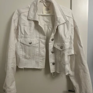 Vit croppad jeansjacka från Stradivarius - Säljer en snygg vit croppad jeansjacka från Stradivarius. Jackan har en cool, rå kant och silverfärgade knappar. Den har långa ärmar och två bröstfickor med lock. Nyskick