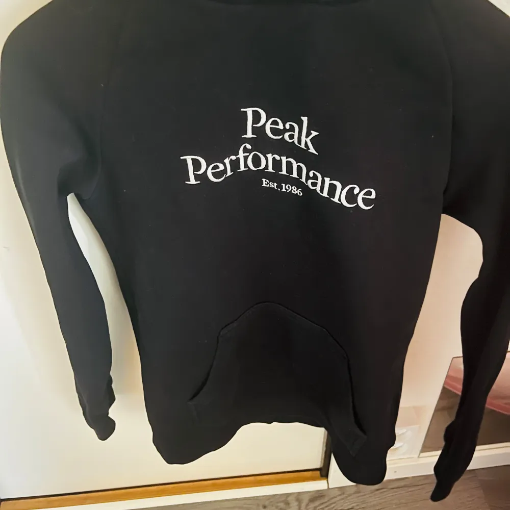 Säljer en svart hoodie från Peak Performance i storlek S. Den är supermjuk och perfekt för kyliga dagar. Klassisk design med vit logga på bröstet och en praktisk känguruficka. Huvan har dragsko för justering. Perfekt för både vardag och chill.. Tröjor & Koftor.