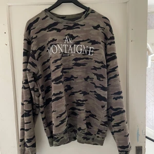 Camouflage tröja från Seven Tees - Säljer en cool camouflage tröja från Seven Tees i storlek XL. Den har ett tryck med texten 'Av. Montaigne' på framsidan och 'LXXV' på baksidan. Perfekt för en avslappnad stil och passar bra till både jeans och joggers. Tröjan är i bra skick och redo för nya äventyr!