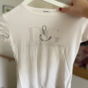 Vit t-shirt från Dolce & Gabbana - Säljer en snygg vit t-shirt från Dolce & Gabbana med glittrande logga framtill. Perfekt för en stilren och lyxig look. T-shirten är kortärmad och i bomull, vilket gör den bekväm och lätt att bära. Passar både till vardags och festliga tillfällen.