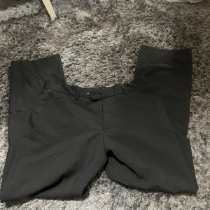 Svarta kostymbyxor - Säljer ett par stiliga svarta kostymbyxor i ullblandning. De har en klassisk design med knappar bak och bälteshällor. Perfekta för både formella tillfällen och kontoret. Materialet är 60% ull, 38% polyester och 2% lycra vilket ger en skön passform.