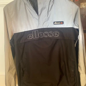 Ellesse  vindjacka 158/164 XXS - Svart/silver jacka med huva och liten dragkedja framtill. Jackan är vatten/vind avvisande. Stor logga på bröstet med en ficka med dragkedja.  I fint skick. Storlek 13/14 år (158/164) Finns i Bandhagen. Kan skickas. Köparen står för frakten.