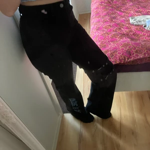 Svets pösiga jeans storlek: S - Svarta jeans som är utsvängda, har spännen på högra benet (detalj) och en blå text på vänstra benet🥰 Sitter tight men samtidigt pösigt på mig! Jag Är runt 160cm lång💕 Skriv ifall du undrar något❤️