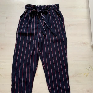 Blårandiga kostymbyxor - Mörkblå kostymbyxor med röda och vita ränder. Resår i midjan. Liten reva på insidan av resåren, men som nyköpta på utsidan. Köpare står för frakt.