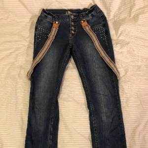 Jeansblå jeans med avtagbara hängslen från KappAhl. Storlek 152. Knappgylf samt tre stycken tryckknappar istället för dragkedja. Strasstenar på framfickorna. Justerbar midja.