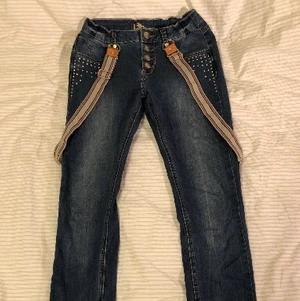 Jeans med hängslen - Jeansblå jeans med avtagbara hängslen från KappAhl. Storlek 152. Knappgylf samt tre stycken tryckknappar istället för dragkedja. Strasstenar på framfickorna. Justerbar midja.