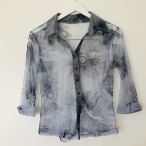 Ljusblå blus med lila knappar  - En fin ljus blå blus med blommönster, lila knappar. Märke okänt, mesh material/ ganska transparant. Så fin och bra skick!