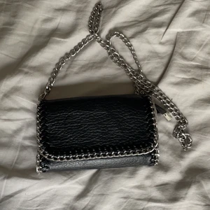 Svart axelremsväska tiamo - Svart axelremsväska i skinnimitation kantad med silverlänk. Ett fack med dragkedja på baksidan och ett fack med dragkedja på insidan. Avtagbar axellänk gör att den kan bäras även som en clutch. Axellänk 105 cm. Längd 21 cm, höjd 14 cm, bredd 5 cm. 💖