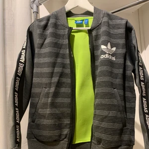 Adidasjacka med dragkedja - En jackliknande kofta som är tunnare än en vanlig jacka men tjockare än en kofta i en bomberjacka modell. I storlek 36 och har detaljer med text ovanpå ärmarna. Grå, svart och vit i speciellt material som håller en varmare. Funkar för träning eller till vardags. Använts endast fåtal gånger.