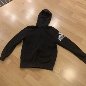Adidas zip-up hoodie - En svart zip-up tröja med ett adidastryck på vänstra ärmen. Dragkedjan är det inget fel på, använt tröjan 3 gånger. Tröjan är storlek 164