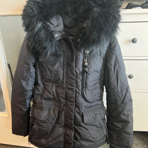 Hollies vinterjacka - Säljer en fin vinterjacka från hollies i storlek 38. Den är hel svart med äkta svart pälskrage. Enda ”felet” på jackan är att den har tappat ”snöret” på dragkedjan som jag visar på bilden. Kostar 4499 på hemsidan just nu