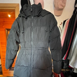 Lång jacka - Helt ny, aldrig använd. Lång svart jacka med luva, dragsko i midjan. Mycket fin och varm!