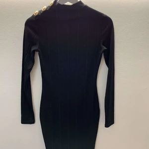 Svart figursydd klänning med guldknappar - Hej! Säljer en Svart figursydd klänning med guldknappar vid ena Axeln. Tyget är ribbad och formars efter dina kurvor. Väldigt snygg och Classy , aldrig använd. Prislapp sitter på. 