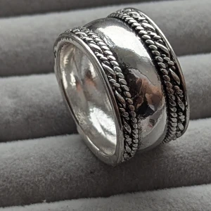 Silverring med flätad design - En bred silverring med en polerad yta och två flätade band som löper längs med ringens kanter. Ringen har en robust och elegant design. Tillverkad av koppar material, nytt