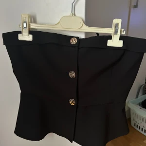 Vero Moda svart topp storlek M - Snygg svart topp från Vero Moda med guldfärgade knappar framtill. Toppen är utan axelband och har en fin volang i nederkanten. Helt oanvänd ny utan prislapp
