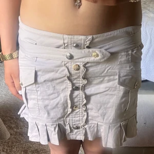 Beige kjol med volangdetaljer - En beige kortkjol med volangdetaljer vid nederkanten. Kjolen har knappar framtill och flera fickor, inklusive en med lock. Den har också en dekorativ bälteffekt i midjan.