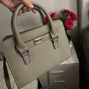 Axelremsväska  - Säljer min fina axelremsväska som är grå med guldiga detaljer. Har en inner dragkedja och två handtag. Man kan ta av sätta på axelremmen.