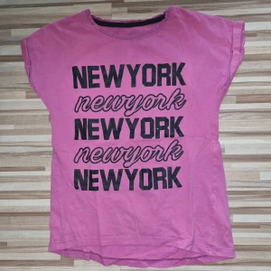 Rosa t-shirt med texten 'NEW YORK' - En rosa t-shirt från Firefly med texten 'NEW YORK' tryckt flera gånger i svart och rosa. T-shirten har korta ärmar och en rund halsringning. Storlek 140