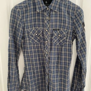 Blå rutig skjorta från Maison Scotch - En blå rutig skjorta från Maison Scotch med långa ärmar och två bröstfickor med knappar. Skjortan har en klassisk krage och knäppning framtill.