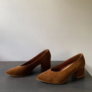 Bruna klackar mocka - Pumps i klassisk modell i mjuk mocka med bred, klädd klack och v-ringad fotöppning. Textilfodrad insida. Klackhöjd ca 5,5 cm i stl 37.  -Slitning på vänster sko framme vid tån -Använda endast en gång -Nypris 499:- 