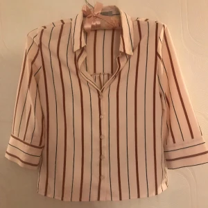 Randig blus  - Rosa blus med ränder. Liknar en skjorta. Knäpps med knappar, längst upp vid halsen är det inga knappar. Är nästan aldrig använd men har dock fått lite bläck från en penna (se sista bilden), annars i fint skick. I materialet polyester och är ganska luftig.