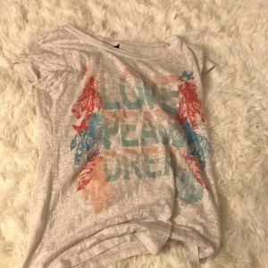 En vit T-shirt - En vit T-shirt där det står i rosa och blåt och de står love peace dreams och har blad runt sig och nån fjäril den är lite genomskinlig så man ser igenom typ
