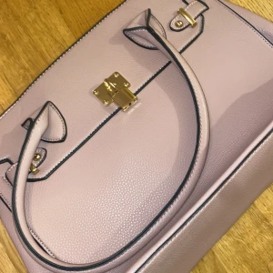 Väska - Babyrosa väska med guldiga detaljer. Väskan är använd några gånger men är i bra skick. Man kan även tajta till väskan med knappen på sidan. 3 fickor inne och en utanför på baksidan. 