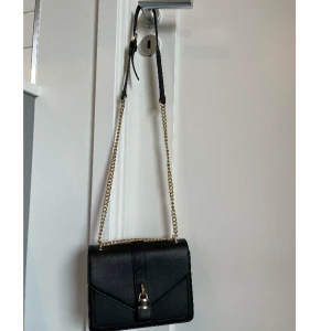 Väska - Svart väska med guldkedjor, hänglås på utsidan som dekoration. Ryms mycket inuti och går att använda bandet som långt eller kort. Tre fack inuti varav ett i mitten med dragkedja. Står inget märke. Nyskick.