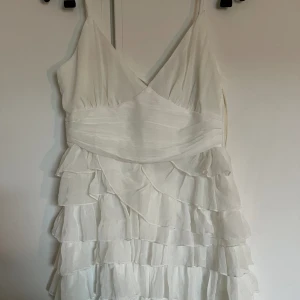 Vit sommarklänning med volanger  - En vit sommarklänning med volanger nertill och fin urringning med tunna axelband som går att justera. I ryggen har den ett knyte som går att dra åt. Aldrig använd, så lapparna sitter kvar.  Från Melissa Berntsens kollektion för Nakd. I stl 38. 