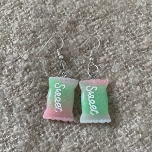 Söta örhängen med godisdesign - Säljer ett par supergulliga örhängen som ser ut som små godispåsar med texten 'Sweet' på. De är i pastellfärger med en mix av grönt, rosa och vitt, och har en glittrig yta.