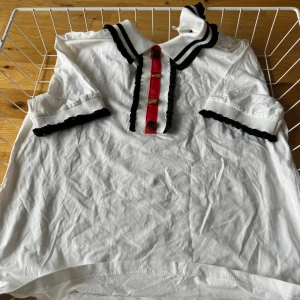 Vit blus med svarta detaljer och röda knappar - Säljer en söt vit blus med svarta detaljer och röda knappar. Blusen har korta ärmar med svarta ränder och en dekorativ röd knappslå framtill. Perfekt för en somrig look eller en casual dag på stan. Blusen är i bra skick och redo för en ny ägare!