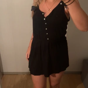 Svart ärmlös klänning med knappar - Säljer en snygg svart ärmlös klänning med knappar framtill. Klänningen har en kort längd och en söt rosett i midjan. Perfekt för sommarens fester eller en utekväll. Den är i nyskick och passar bra för både vardag och fest.