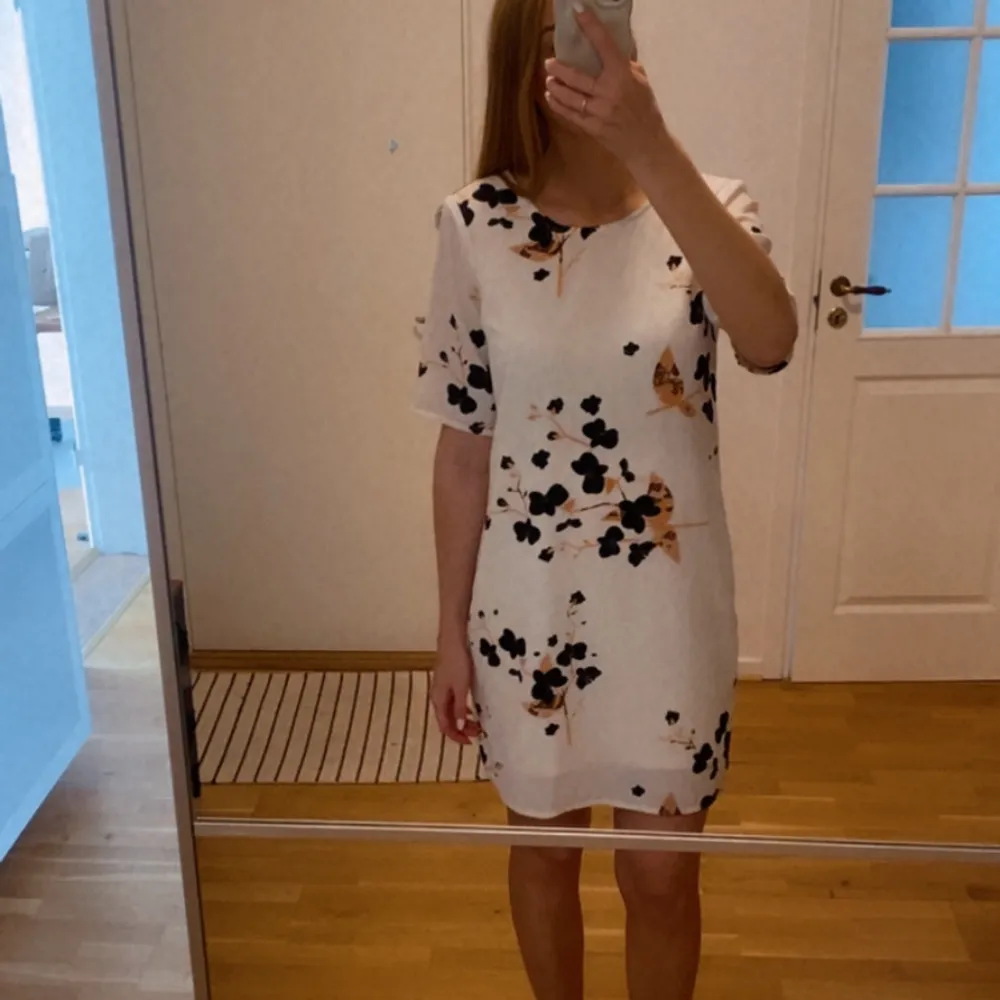 En vit kortärmad klänning med blommigt mönster i svart och rosa. Klänningen har en rund hals och en öppen ryggdetalj. Snodden som stänger knappen bak har torkat ut och behöver eventuellt bytas. Annars som ny.. Mekot.