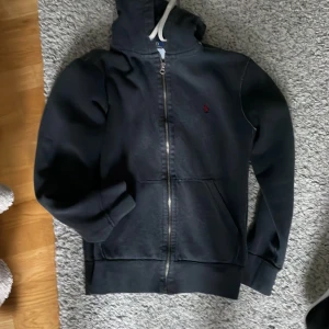 Svart hoodie från Polo Ralph Lauren - Säljer en svart hoodie från Polo Ralph Lauren i storlek S. Den har en dragkedja framtill och en liten röd logga på bröstet. Hoodien är i bra skick och perfekt för höst och vinter. Den har en mysig insida och är superbekväm att ha på sig. Passar både till vardags och träning!