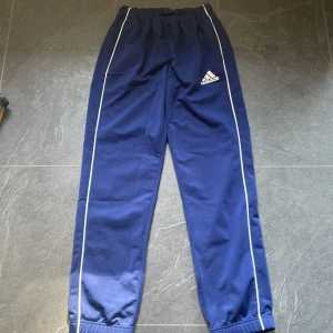 Blå träningsbyxor från Adidas - Säljer ett par blå träningsbyxor från Adidas. De har en elastisk midja och vita detaljer längs benen. Byxorna är i bra skick och perfekta för träning eller en avslappnad dag. Adidas-loggan är broderad på framsidan.