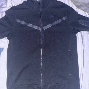 Nike tech svart - Säljer en svart zip hoodie från Nike i storlek XL. Jackan har en dragkedja framtill och en snygg design med diskreta detaljer på ärmarna. Perfekt för träning eller vardagsbruk. Den är i bra skick och redo för en ny ägare!