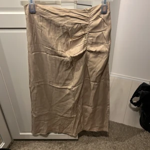 Beige kjol från Kappahl - En beige kjol från Kappahl med rynkade detaljer på framsidan. Kjolen har en slits på sidan och är tillverkad i ett glansigt material.