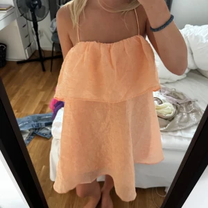 Persikofärgad klänning - En persikofärgad kortklänning med volangdetaljer. Klänningen är ärmlös och har smala axelband som korsas i ryggen. Denna klänning kan man välja själv om man vill ha banden uppe eller lägga ner dem!!❤️❤️