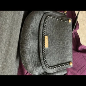 Svart axelväska från Cosmopolitan - En elegant svart axelväska från Cosmopolitan. Väskan har en texturerad yta och en framficka med dekorativa sömmar och guldfärgade nitar. Den har en justerbar axelrem och en liten metallplatta med märkesnamnet på framsidan.