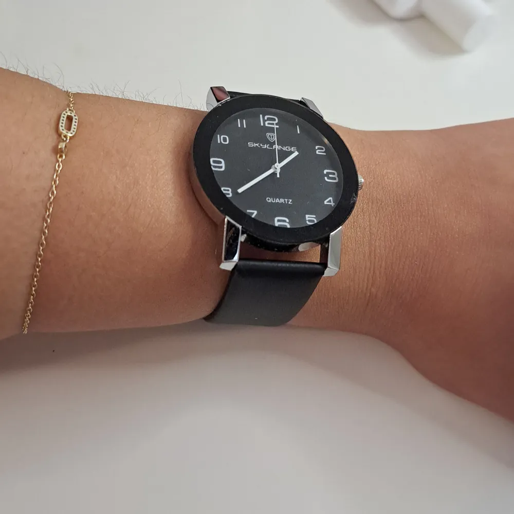 En elegant svart klocka från Skylange med en rund urtavla och tydliga vita siffror. Klockan har en svart rem och en silverfärgad spänne. Den är av quartz-typ och har en minimalistisk design.. Accessoarer.