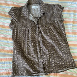 Brun blus med blommönster från Sise - En brun blus med ett blommönster i beige och gula detaljer från märket Dice. Blusen har korta ärmar och knappar framtill. 