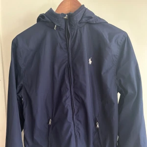 Raulph Lauren Jacka - Snygg Raulph Lauren jacka som passar både sommar och höst. Använd ett fåtal gånger så den är i nyskick. Skönt material och fin blå färg. Luvan är avtagbar. Nypris Kids Brandstore 2100:- Stl 170  Passar både killar & tjejer