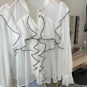 Oanvänd blus från Zara! - Storleken är Xs men stor i storleken! Jättefin vit blus med svarta detaljer samt volanger! Den är lite genomskinlig men går bra att ha ett linne under! Använt endast cirka 3 gånger 